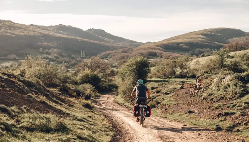 Ciclista pedaleando por un camino rural en España, disfrutando de sus viajes en bicicleta por España. Paisaje montañoso y vegetación verde.