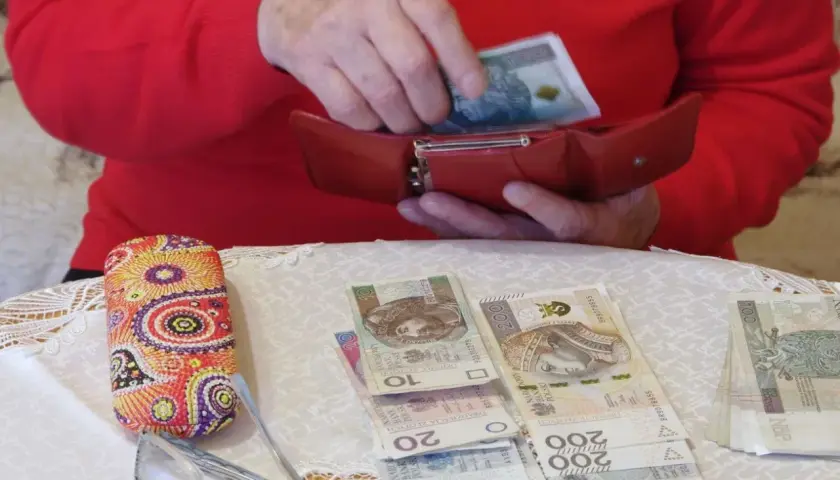 Dłonie chowają banknot 100 zł do czerwonego portfela. Obok leżą pieniądze, okulary i etui. To pokazuje, gdzie trzymać pieniądze.