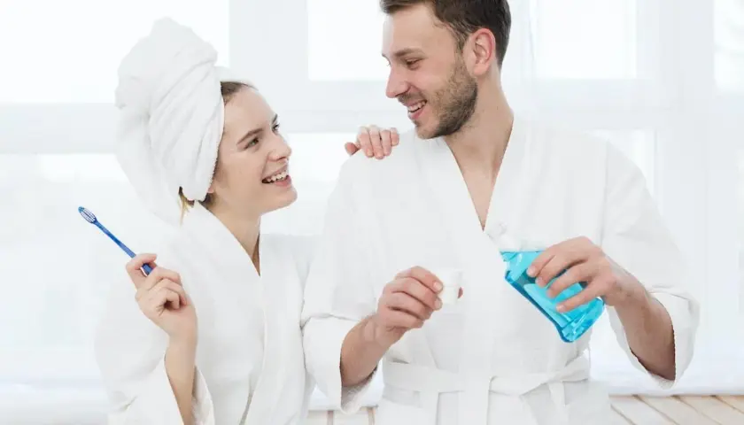 Coppia sorridente in accappatoio, lei con spazzolino, lui versa collutorio. Imparare come usare il collutorio per un sorriso smagliante.