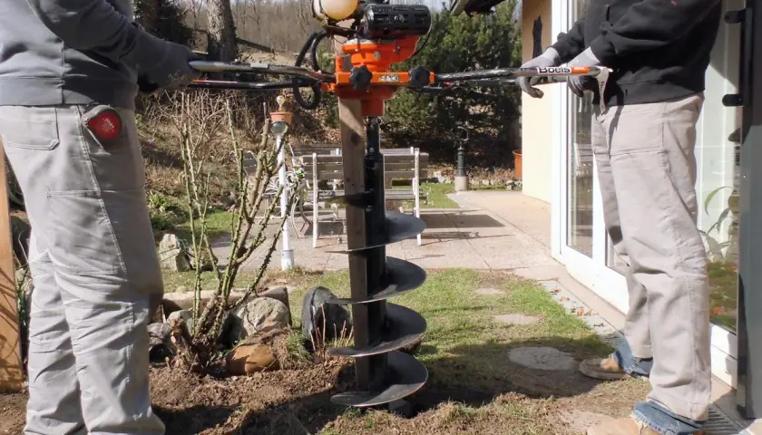 Zwei Personen bedienen einen leistungsstarken **Erdhandbohrer** im Garten. Das Gerät mit seinem orangefarbenen Motor und der langen Bohrschnecke ist bereit, Löcher für Pfosten oder Pflanzen zu graben.