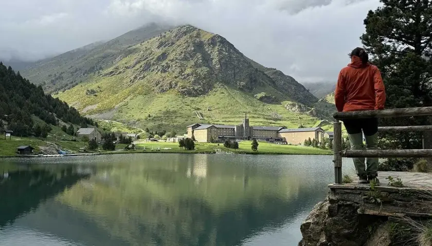 Persona contempla el sereno lago en el valle de Nuria, con imponentes montañas verdes y un complejo de edificios al fondo.