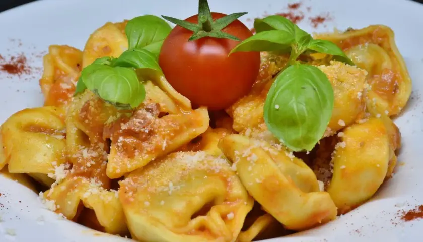 Pyszne tortellini na talerzu, polane sosem pomidorowym, ozdobione listkami bazylii i pomidorkiem.