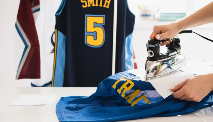 Une main utilise un fer à repasser pour défroisser un maillot de basket bleu avec des lettres jaunes. D'autres maillots sont suspendus en arrière-plan.