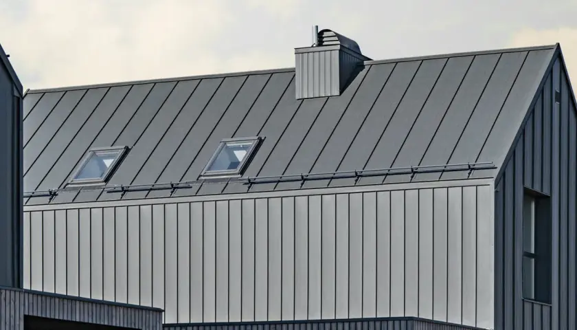Nowoczesny budynek z szarą blachą na rabek, dwoma oknami dachowymi i kominem.