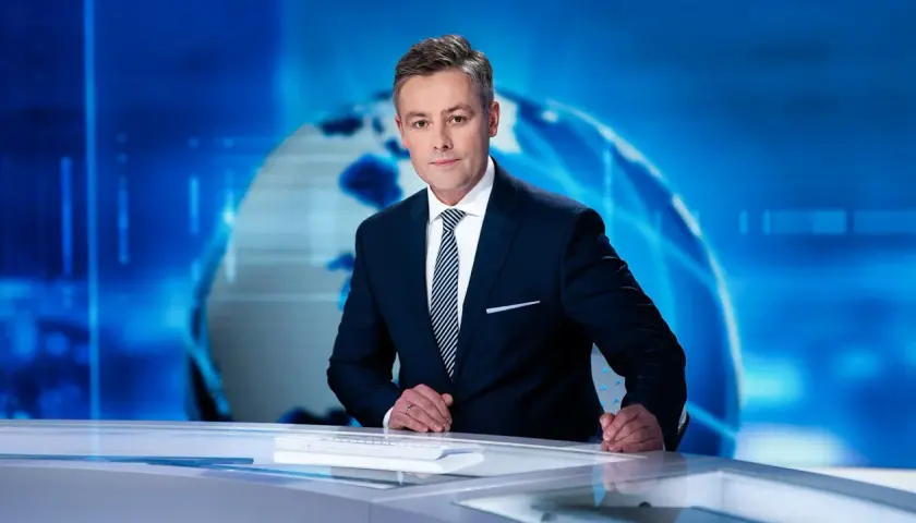 Elegancki konferansjer w garniturze stoi przy biurku na tle niebieskiej grafiki z globusem.