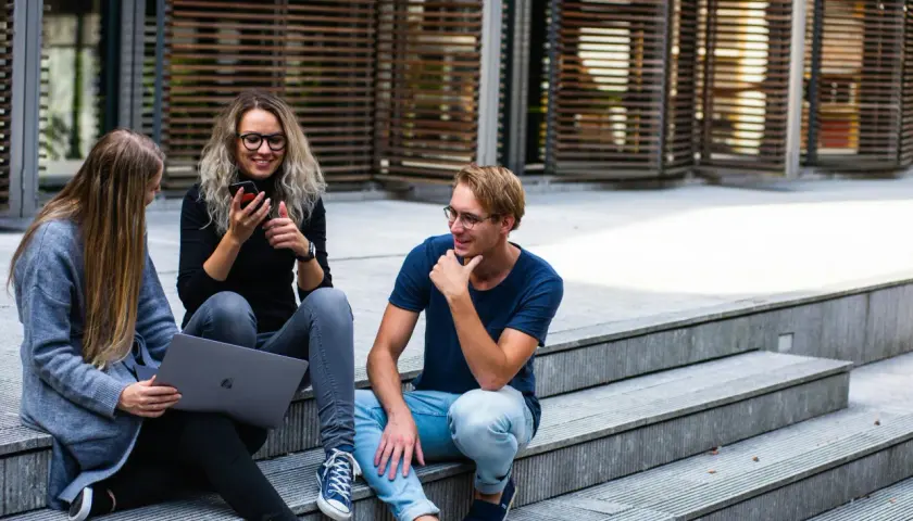 Drei junge Leute sitzen auf einer Treppe. Eine Frau mit Laptop und eine Freundin mit Handy, die sich über das Studium zuhause unterhalten.
