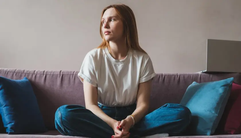 A young woman with red hair sits cross-legged on a couch, looking away thoughtfully.