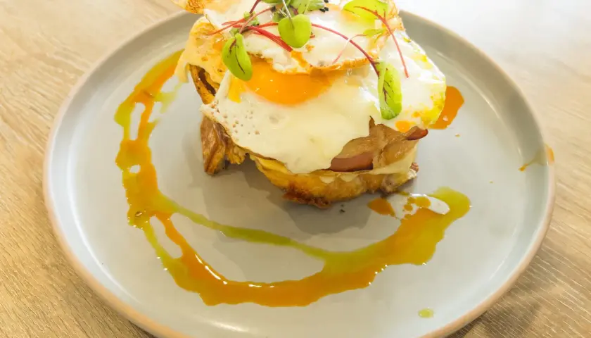 Un délicieux croque madame avec un œuf au plat coulant, du jambon et du fromage fondu, garni de jeunes pousses et servi sur une assiette avec un filet de sauce.