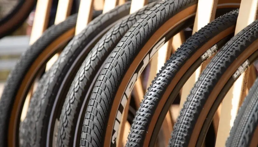 A row of diverse bicycle tires, featuring knobby mountain bike tires and smoother road bike tires, all with distinctive tan sidewalls, are displayed.