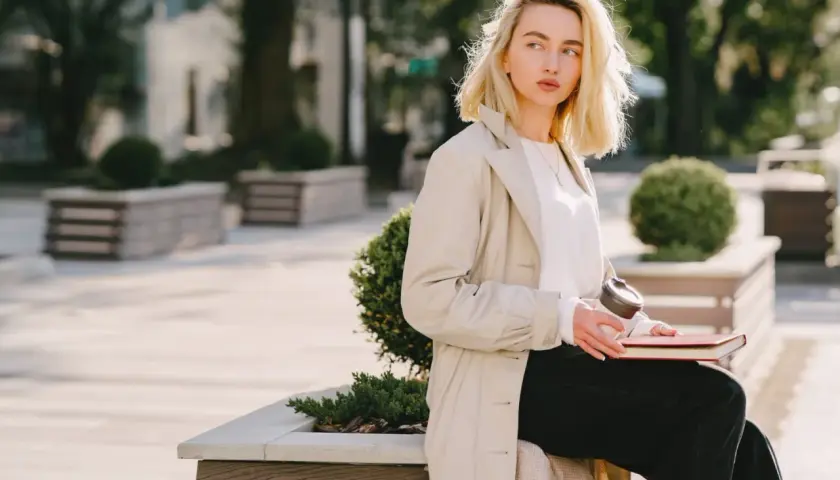 Une femme blonde, assise dehors avec un livre et un café, porte un trench beige et un haut blanc. Parfait pour savoir comment s'habiller quand il fait 15 degrés.