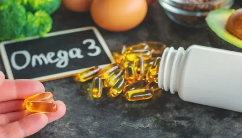 Kapsułki Omega-3 wysypujące się z białej butelki, na tle tabliczki z napisem "Omega 3".