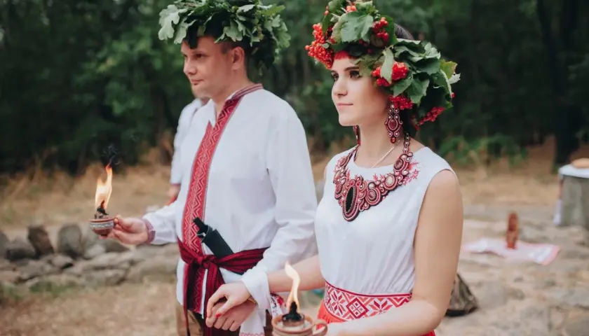 Para młoda w tradycyjnych strojach, z wiankami na głowach, trzymająca płonące świece. To piękna swadźba.