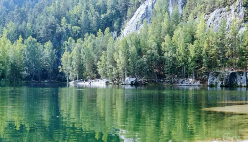 Zielone jezioro otoczone lasem i skalnymi formacjami. W oddali widać ludzi spacerujących po plaży. To malownicze miejsce, gdzie góry stołowe spotykają się z wodą.