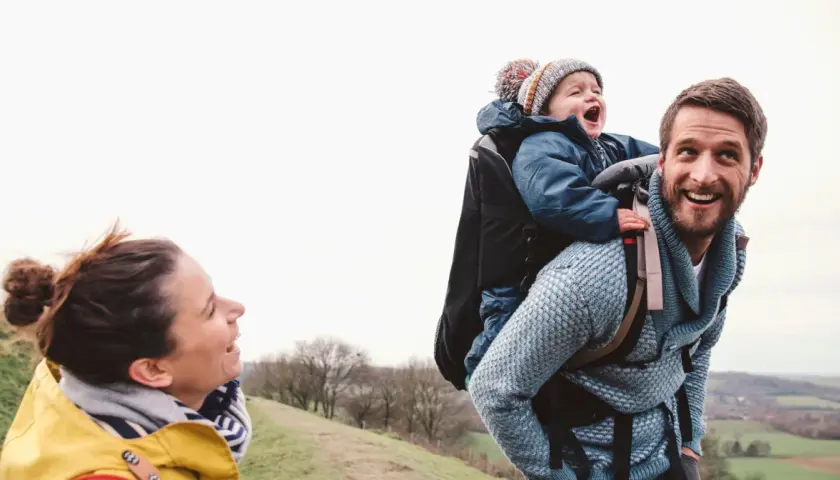 Szczęśliwa rodzina na spacerze. Tata niesie dziecko w nosidełku turystycznym, mama uśmiecha się do nich.