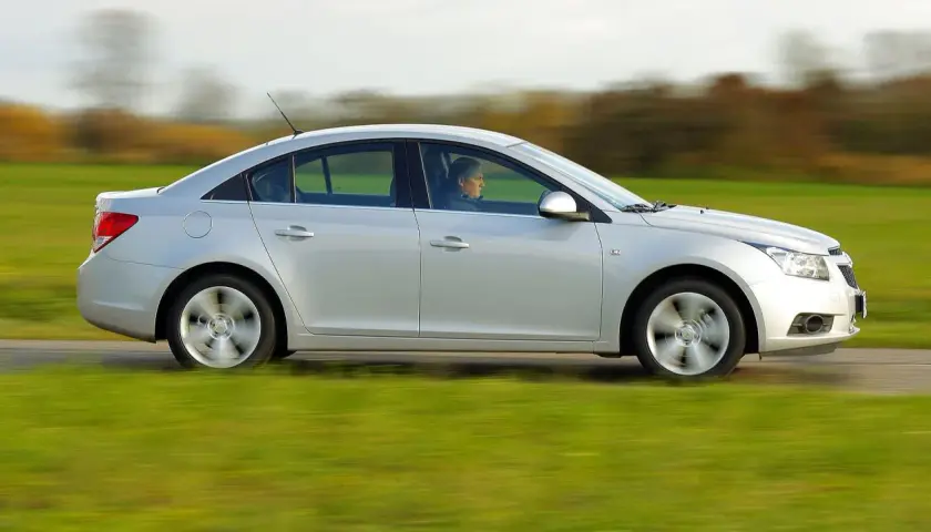 Srebrny Chevrolet Cruze 1.8 benzyna w ruchu, z kierowcą za kierownicą, przemierza wiejską drogę pośród zielonych pól.
