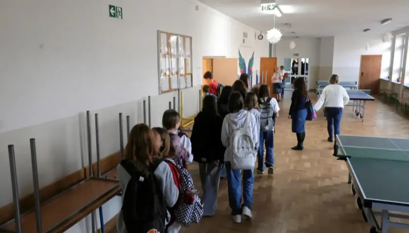 Uczniowie w kolejce na szkolnym korytarzu, w tle stoły do tenisa stołowego.