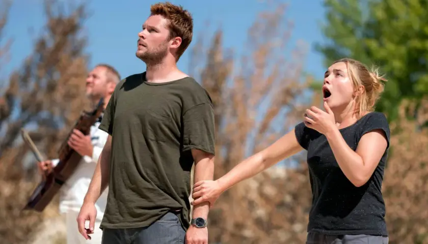 En kvinna griper tag i en mans arm, båda ser uppåt med förvånade uttryck. En man med ett instrument syns i bakgrunden. Scen från Midsommar filmens rollista.