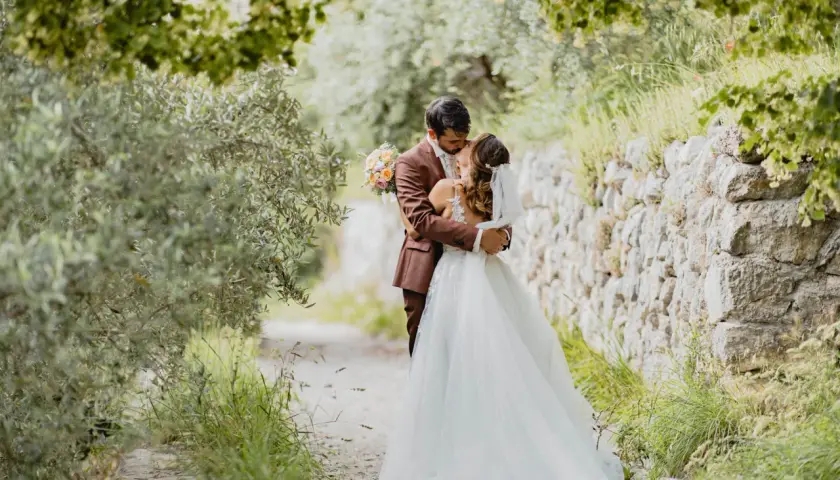 Un couple s'embrasse tendrement lors de leur mariage de 50 personnes, entouré par la nature verdoyante et un mur de pierres anciennes.
