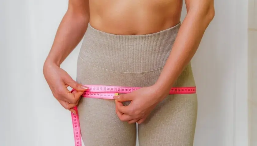 Mujer midiendo su cintura, observando la cinta métrica rosa que rodea sus caderas, enfocándose en los contornos y posibles hip dips.