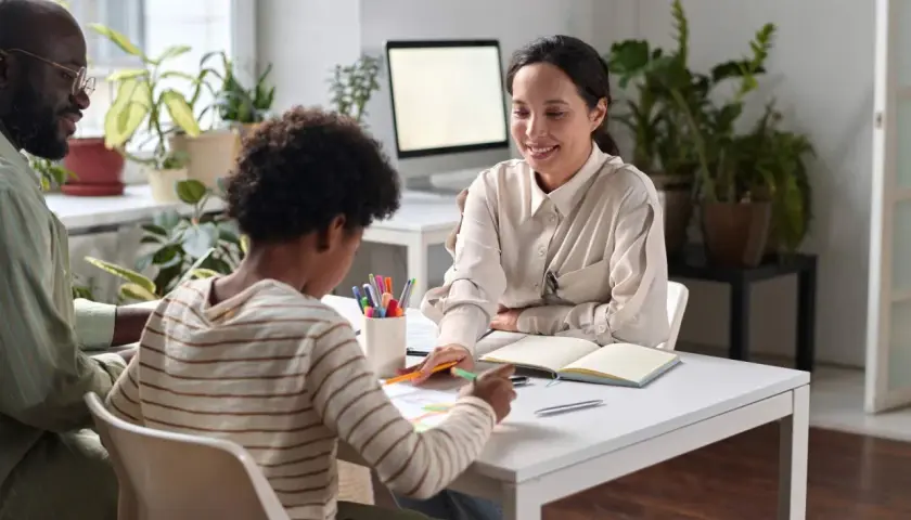 Ein Vater, sein Kind und eine freundliche Frau sitzen an einem Tisch. Die Frau, die wie eine Integrationshelferin aussieht, erklärt etwas. Das Kind malt. Das Gehalt eines Integrationshelfers ist eine wichtige Frage für viele.