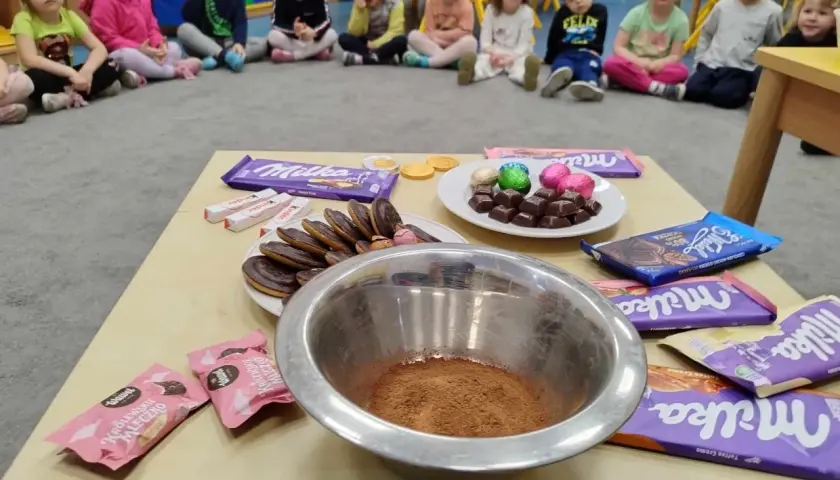 Dzieci w przedszkolu świętują dzień czekolady. Na stole czekolady Milka, Kinder, ciastka i miska z kakao.