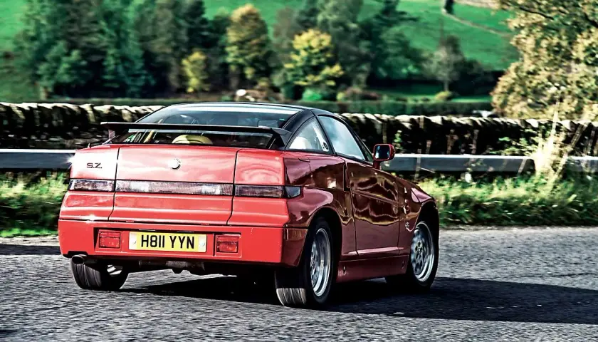 Czerwone Alfa Romeo SZ z czarnym dachem i spoilerem jedzie po wiejskiej drodze.