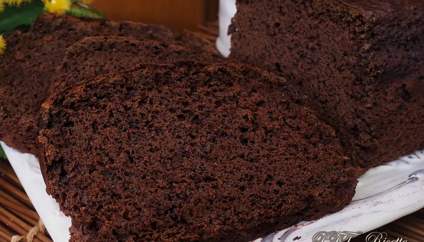 Plumcake al cacao sofficissimo, affettato su un piatto bianco, con fiori gialli sullo sfondo.