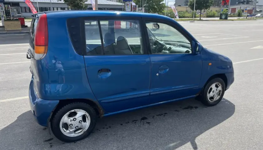 En blå Hyundai Atos står parkerad vid en bensinstation. Bilen har några rostfläckar.