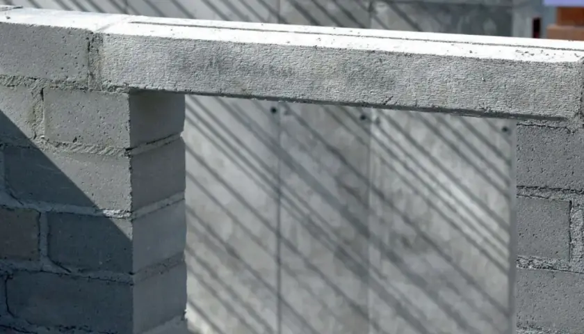 Betonowy nadproże nad otworem w budowie, podparty na bloczkach. Cienie ukośnie przecinają tło.