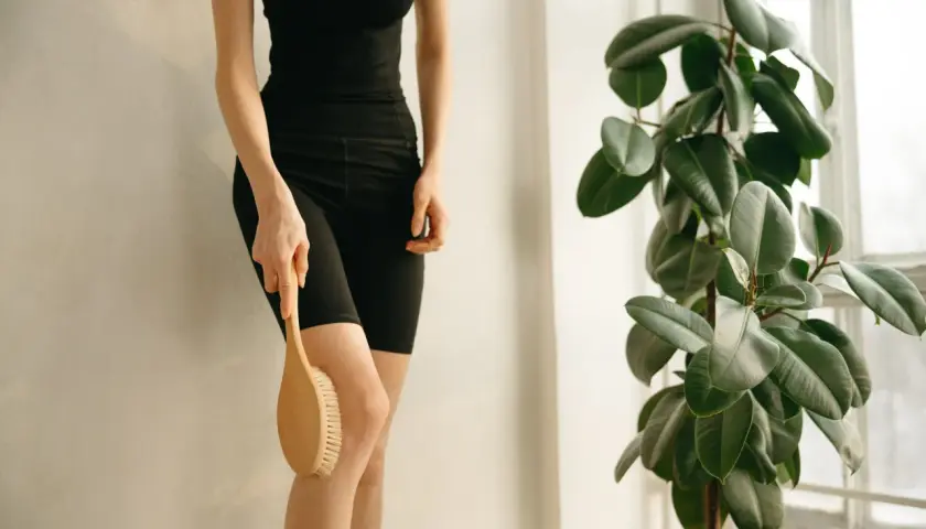 En person utför lymfmassage hemma med en borste på benet, bredvid en stor grön växt.