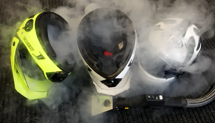 Pulire interno casco moto con vapore. Tre caschi, uno giallo fluo, uno bianco e nero, uno bianco, circondati da fumo.