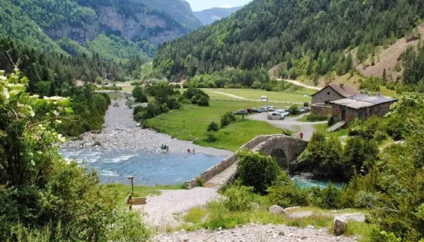 Un río de aguas turquesas fluye por un valle verde, cruzado por un puente de piedra. La ruta Bujaruelo invita a disfrutar de este paisaje montañoso.