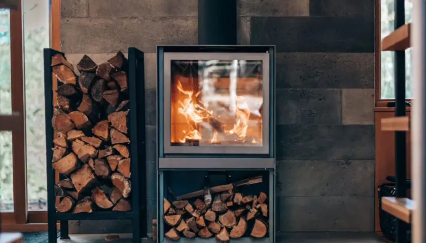Płonąca koza na tle kamiennej ściany. Obok stojak z drewnem, a pod piecem schowek na opał.