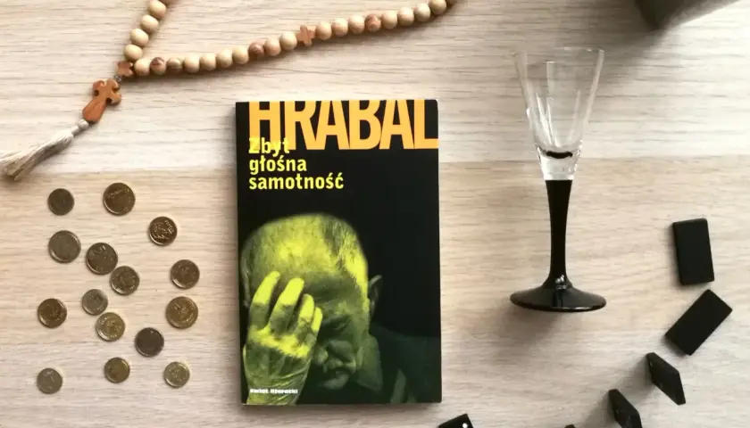 Koraliki różańca, monety, kieliszek i domino otaczają książkę "Zbyt głośna samotność".