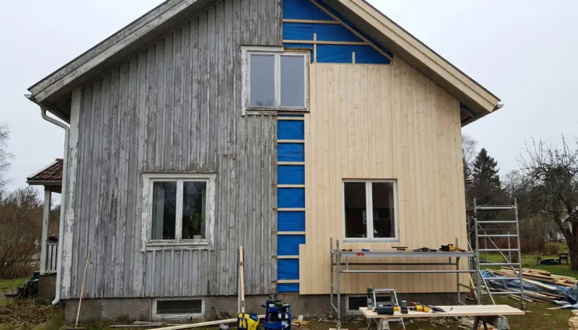 Husfasaden byts ut. Gamla träpanelen på vänster sida kontrasterar mot den nya, ljusa panelen som monteras på höger sida.