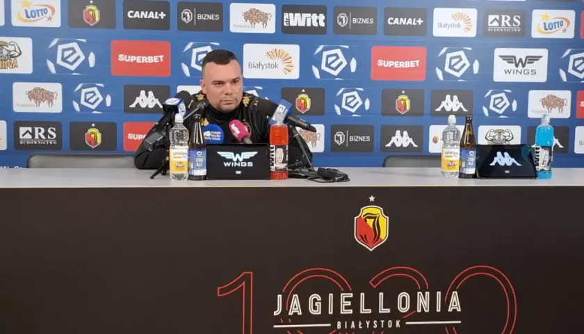 Mężczyzna w czarnym stroju siedzi przy stole konferencyjnym, na którym stoją napoje. Tło zdobią logotypy sponsorów, w tym Jagiellonia Białystok, co sugeruje kontekst związany z jagiellonia transfery.