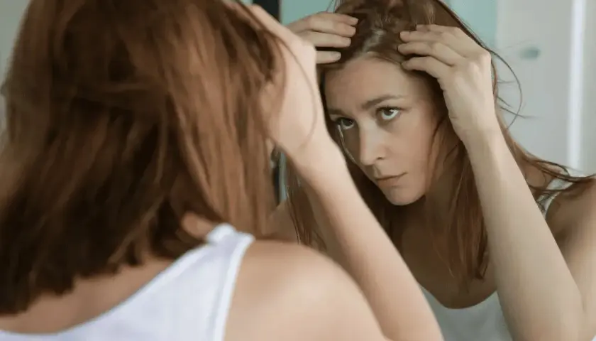 Mujer preocupada revisa su cuero cabelludo en el espejo, buscando soluciones sobre como frenar la caida del cabello.