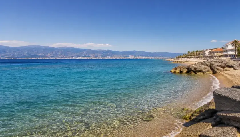 Spiaggia di Reggio Calabria con mare cristallino, palme e case bianche. All'orizzonte, montagne azzurre sotto un cielo limpido.