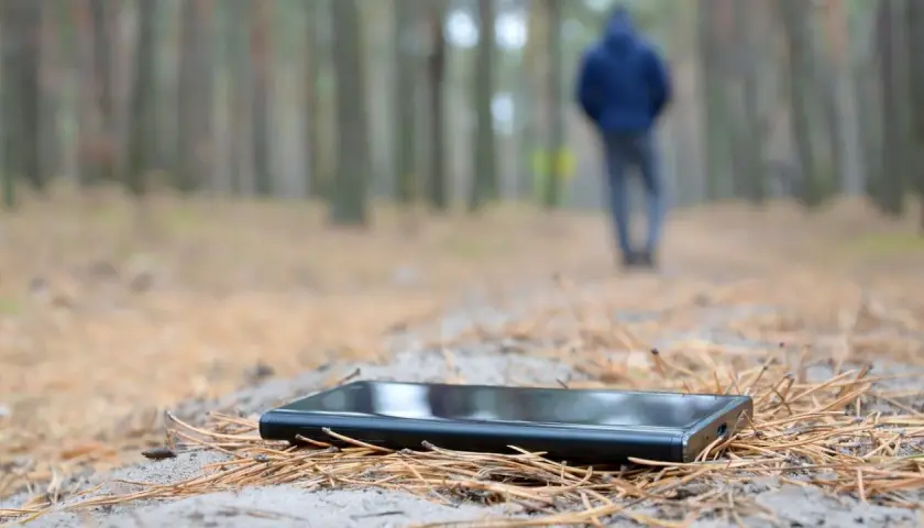 Ein Handy verloren, was tun? Ein schwarzes Smartphone liegt im Wald auf Nadeln. Im Hintergrund geht eine Person weg.