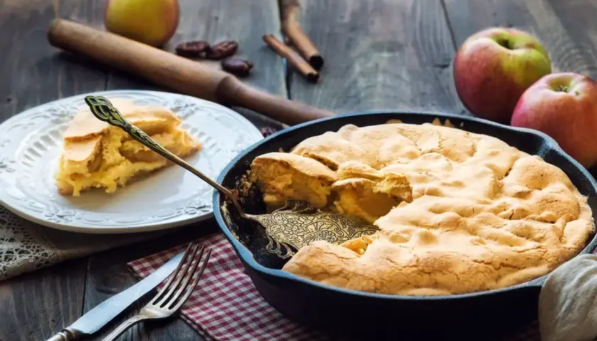 Una deliziosa torta di mele in padella, appena sfornata, con una fetta servita su un piatto. Mele fresche e cannella completano la scena rustica.