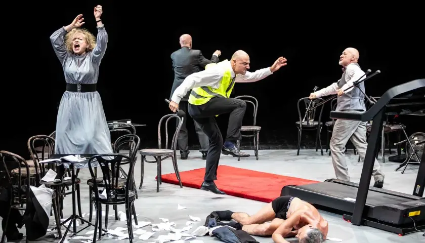 Scena teatralna z kobietą w szarej sukni, mężczyzną w kamizelce odblaskowej i leżącym na bieżni. Czy to symbol tego, ile trwają przerwy w teatrze?