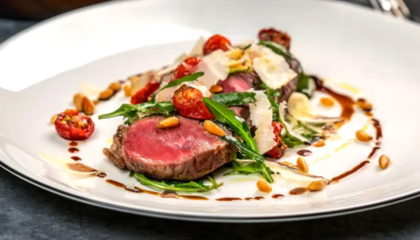Saftiges Tagliata di Manzo mit Rucola, Kirschtomaten, Pinienkernen und Parmesan auf einem weißen Teller.