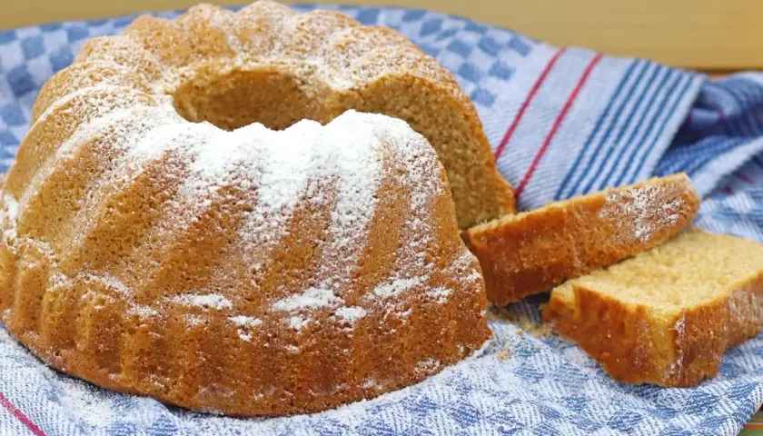 Puszysta babka panettone, oprószona cukrem pudrem, leży na niebiesko-białym ręczniku. Obok niej leżą dwa kawałki ciasta.