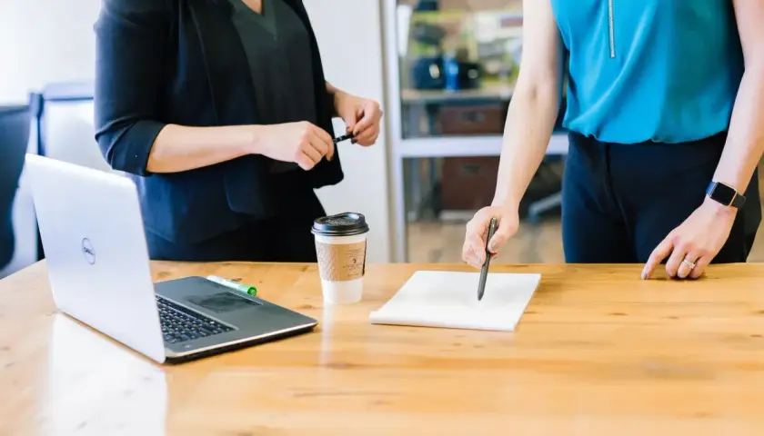 Zwei Frauen besprechen Dokumente am Tisch. Ein Laptop und Kaffee stehen bereit. Möglicherweise geht es um die Auszahlung Lebensversicherung Steuer.