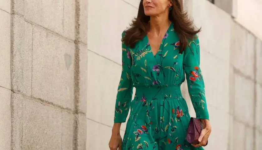 La reina Letizia, con un vestido verde estampado y pendientes llamativos, camina con elegancia junto a un muro de piedra.