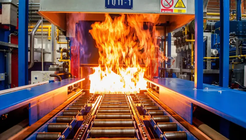 Płomienie buchają z pieca, w którym odbywa się nawęglanie stali. Taśmociąg z rolkami transportuje metal przez gorący proces.