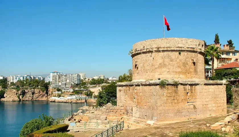 Antalya Altstadt: Ein runder Steinturm mit türkischer Flagge überragt die Bucht. Im Hintergrund moderne Gebäude und Felsen.