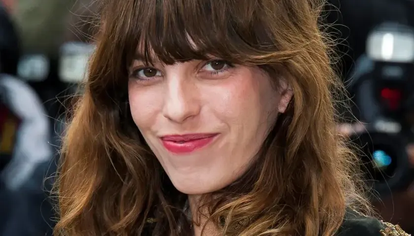 Kobieta z brązowymi włosami i charakterystyczną grzywką w stylu french bangs uśmiecha się lekko.