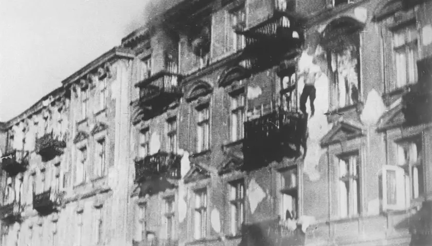 Brennende Gebäude im Warschauer Ghetto. Rauch steigt auf, Fenster sind zerbrochen, Balkone sind dunkel.