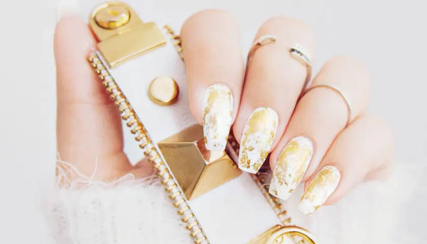 En hand med vita naglar dekorerade med guld nagellack, som en lyxig touch. En vit och guldig accessoar syns också.